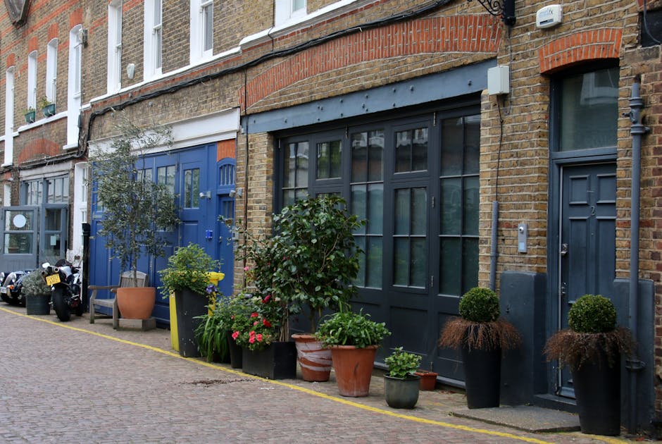 A person wearing a blue jacket and patterned shirt is using hedge trimmers to trim a leafy hedge along the side of a garden. The garden features a wooden table with potted plants, a small black contai